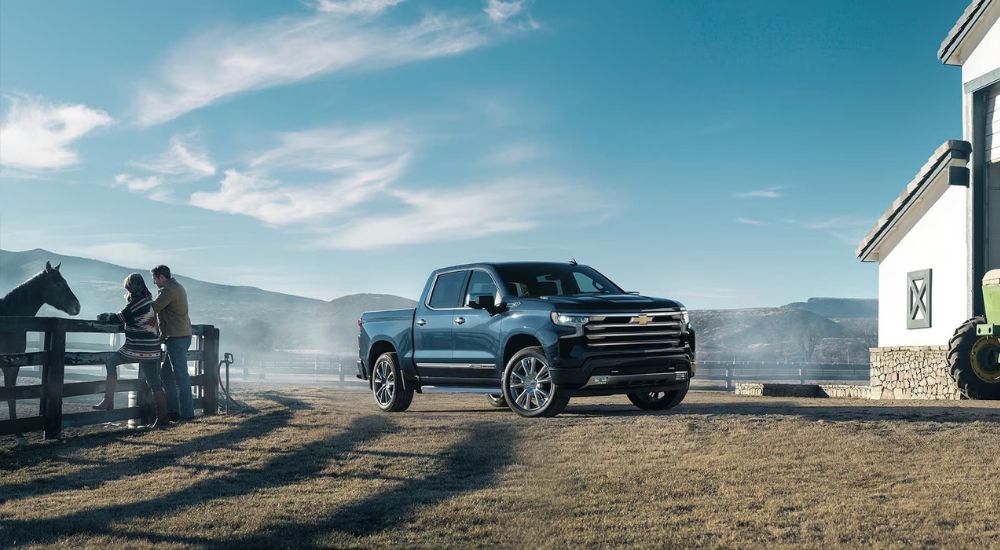 A blue 2024 Chevy Silverado 1500 High Country is shown parked near a wooden fence.