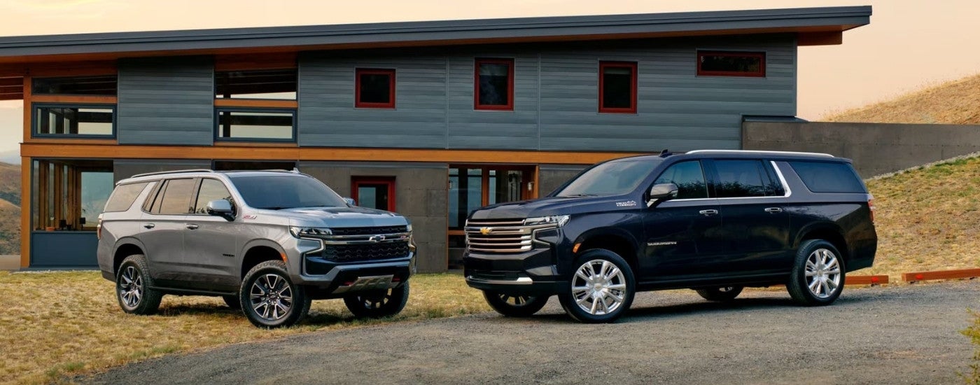 Two 2022 Chevy SUVs, a grey Chevy Tahoe Z71 and a dark blue Suburban High Country, are shown in front of a house.