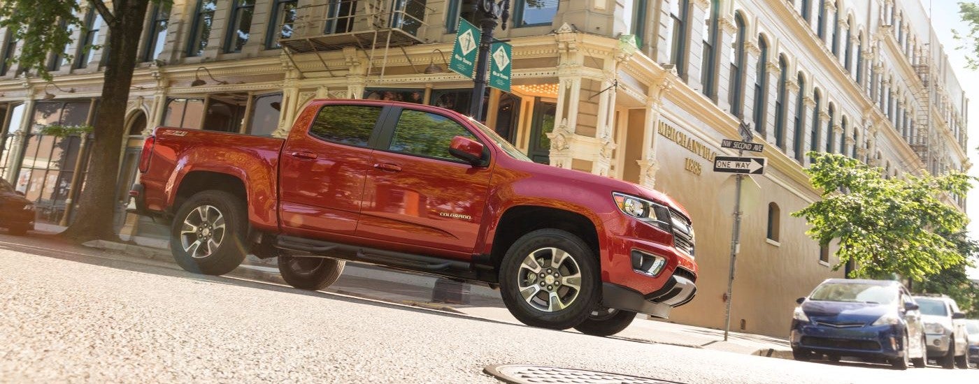 An older Chevy truck, a red 2015 Chevy Colorado, is shown from a low angle driving on a city street.