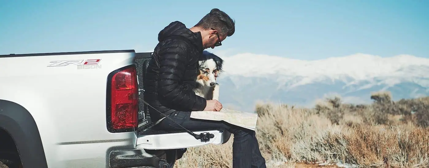 A man and his dog sit on the tailgate of a white 2019 Chevy Colorado as they look at a map.